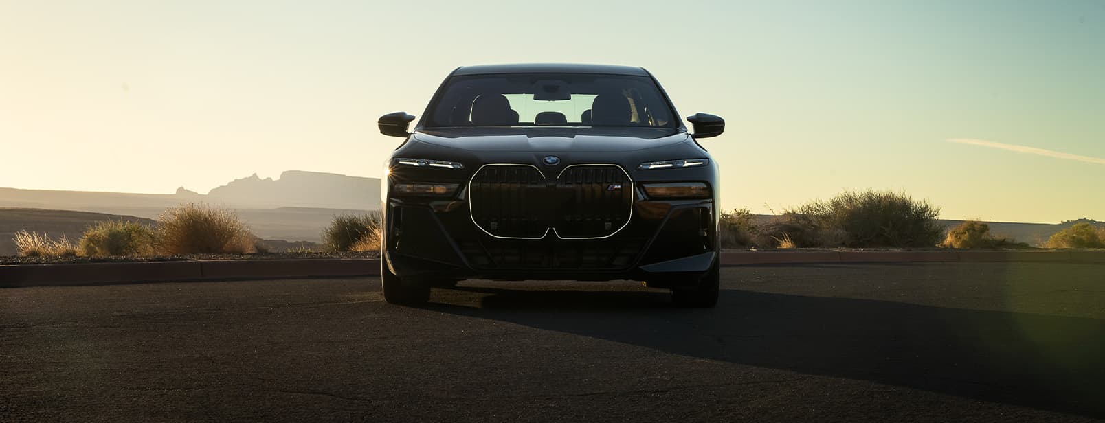 Front profile of a black 2026 BMW i7 M70 parked in flat, desert landscape with the sun setting in the background.