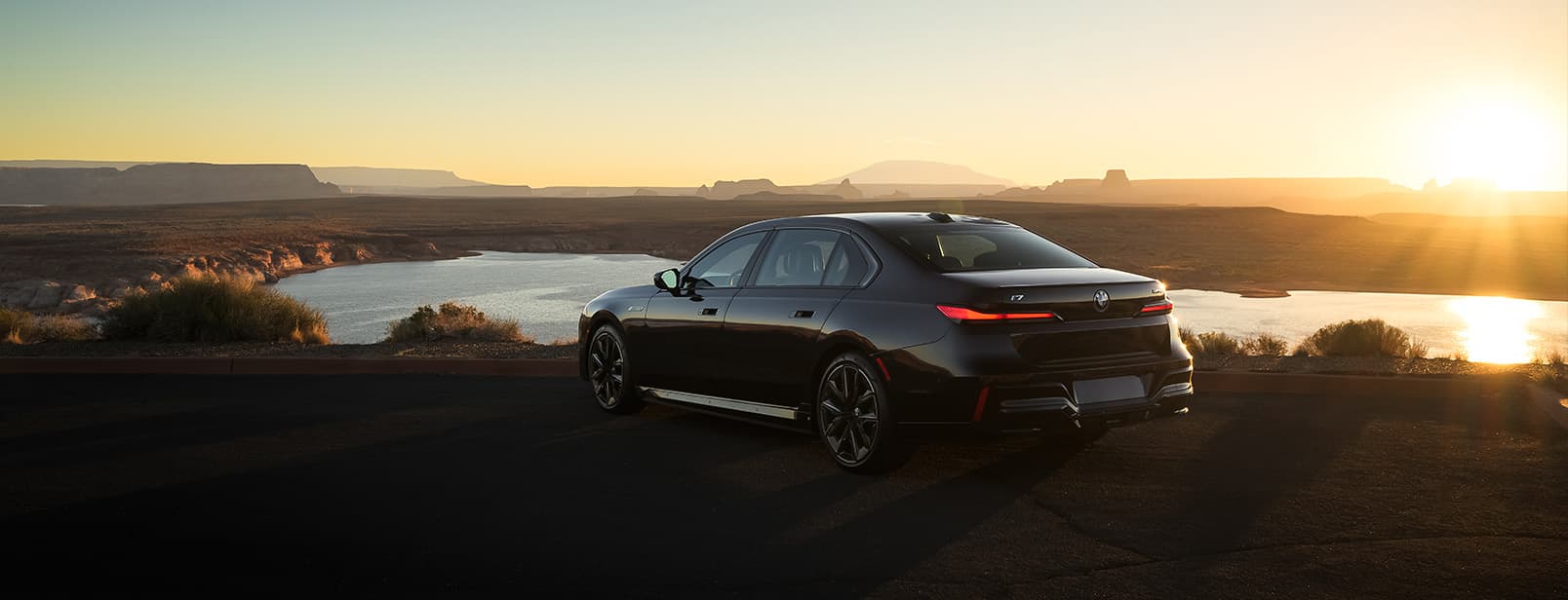 Rear and side profile of a black 2026 BMW i7 M70 parked in flat, desert landscape with the sun setting in the background.