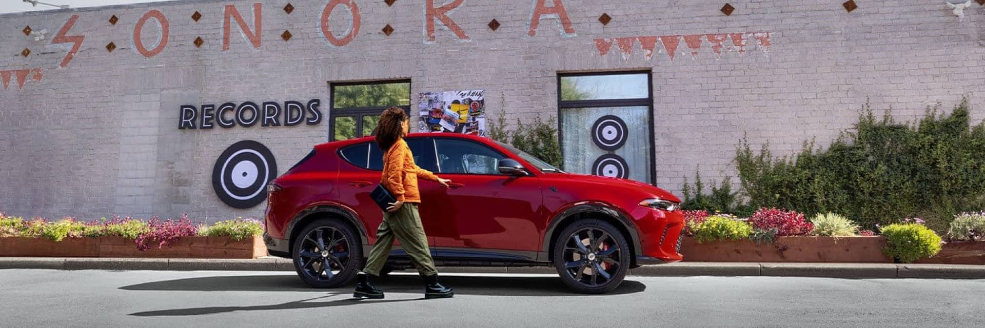 a person walking toward the passenger side door of a red Dodge Hornet in front of a record store