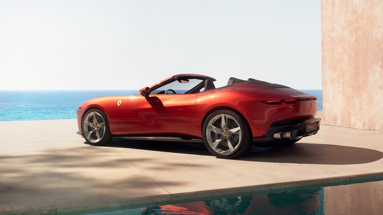 Rear three-quarter view of a red Ferrari convertible parked by the ocean on a sunny day.