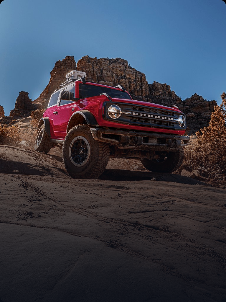 Red Ford Bronco climbing over rocky desert terrain with sandstone formations in the background.