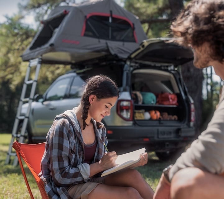  Ford Bronco Sport with a camping tent on the roof and woman sits in chair in front reading 