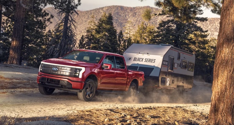 A red Ford F-150 towing a trailer on a road near mountains.