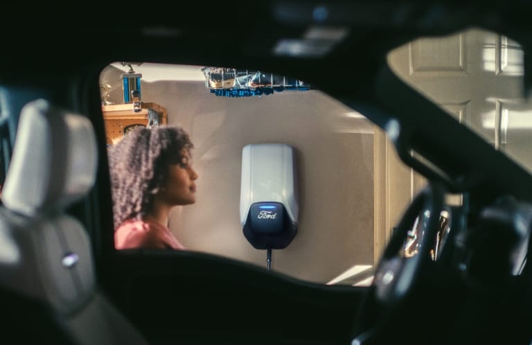 A Ford Connected Charge Station mounted on a garage wall with a woman seen through a Mustang Mach-e window.