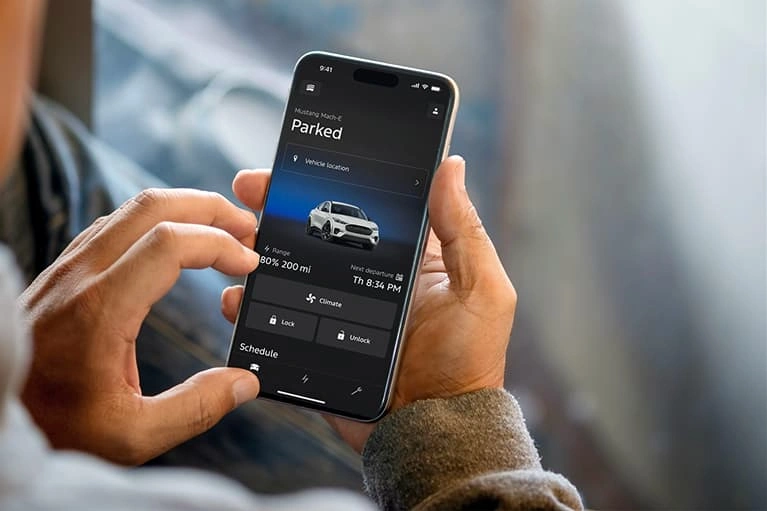 Person holding a smartphone displaying the Ford Pass app showing a parked Mustang Mach-E with battery range and vehicle controls.