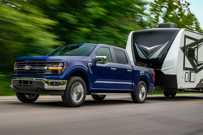 Blue Ford F-150 towing a large travel trailer on a tree-lined highway.