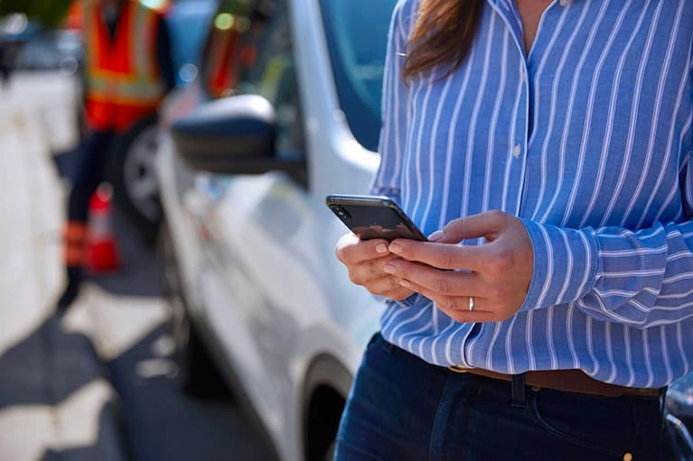 Woman using her phone roadside next to a vehicle with a roadside assistance worker in the background.