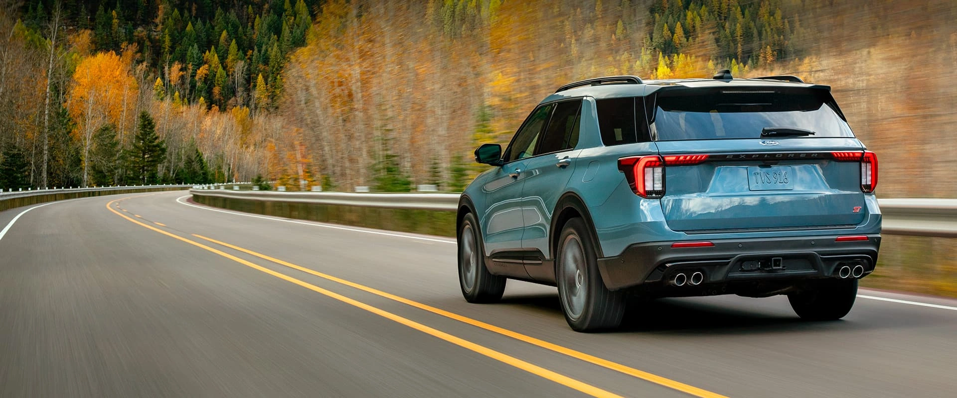 Rear view of a blue Ford Explorer ST driving on a winding road surrounded by fall foliage.