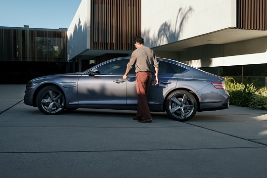 Driver side profile view of driver reaching for door handle on Genesis G80 parked beside modern home