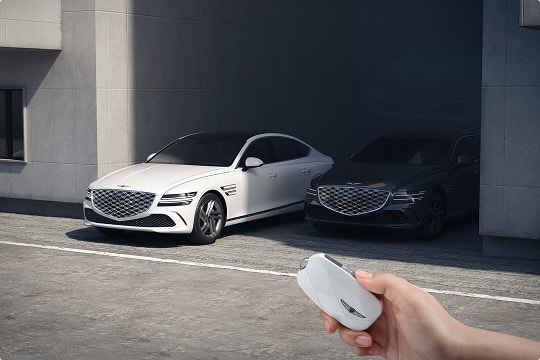 Genesis G80 in Uyuni White parked beside black G80, remote key in foreground near modern garage