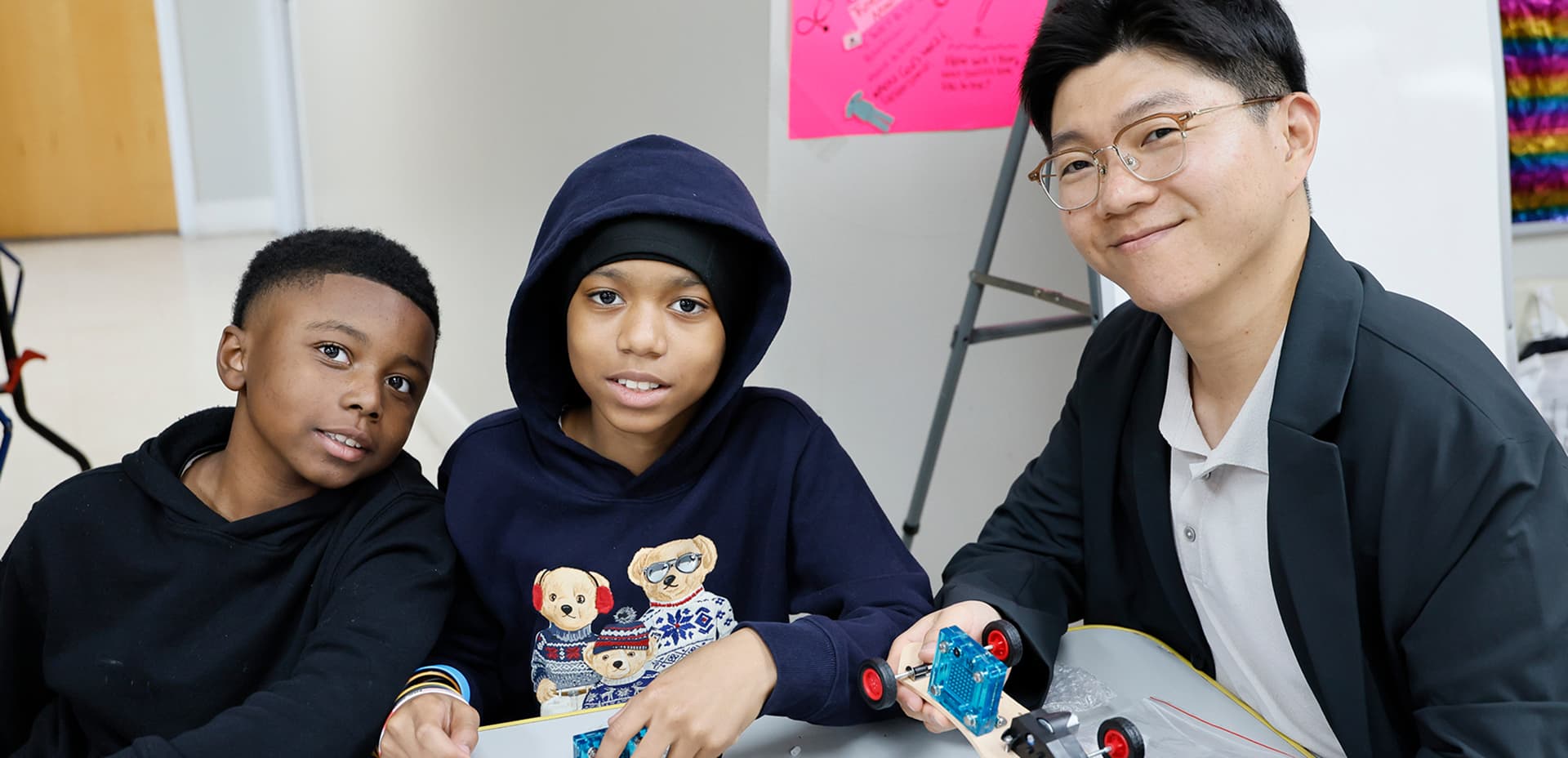 Two students and an adult mentor working together on a small wheeled robotics project in a classroom.