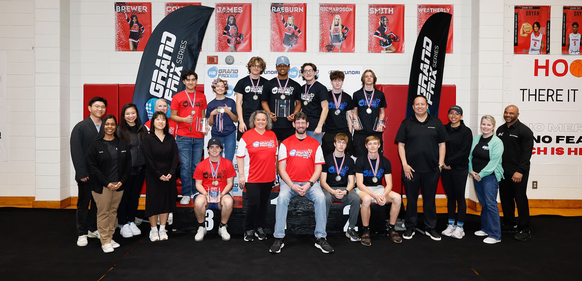 Group of students and adults posing on a winner's podium at a Hyundai H2 Grand Prix Series competition in a school gymnasium.