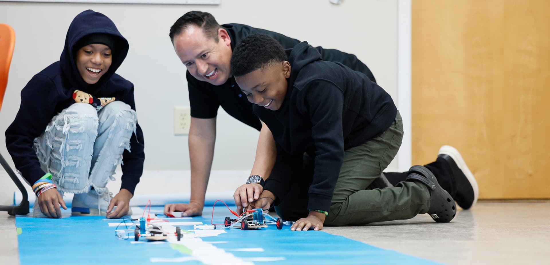 An adult and two children smiling while testing a small hydrogen-powered toy car on the floor during a STEM activity.