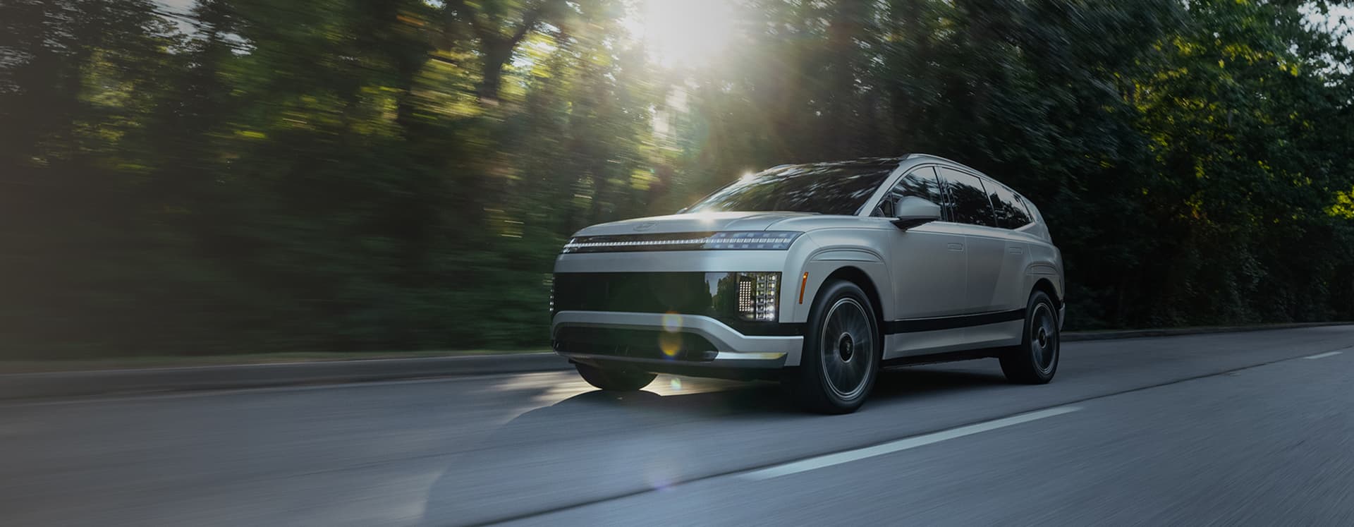 White electric SUV driving on a tree-lined road with motion blur and sunlight breaking through the forest.
