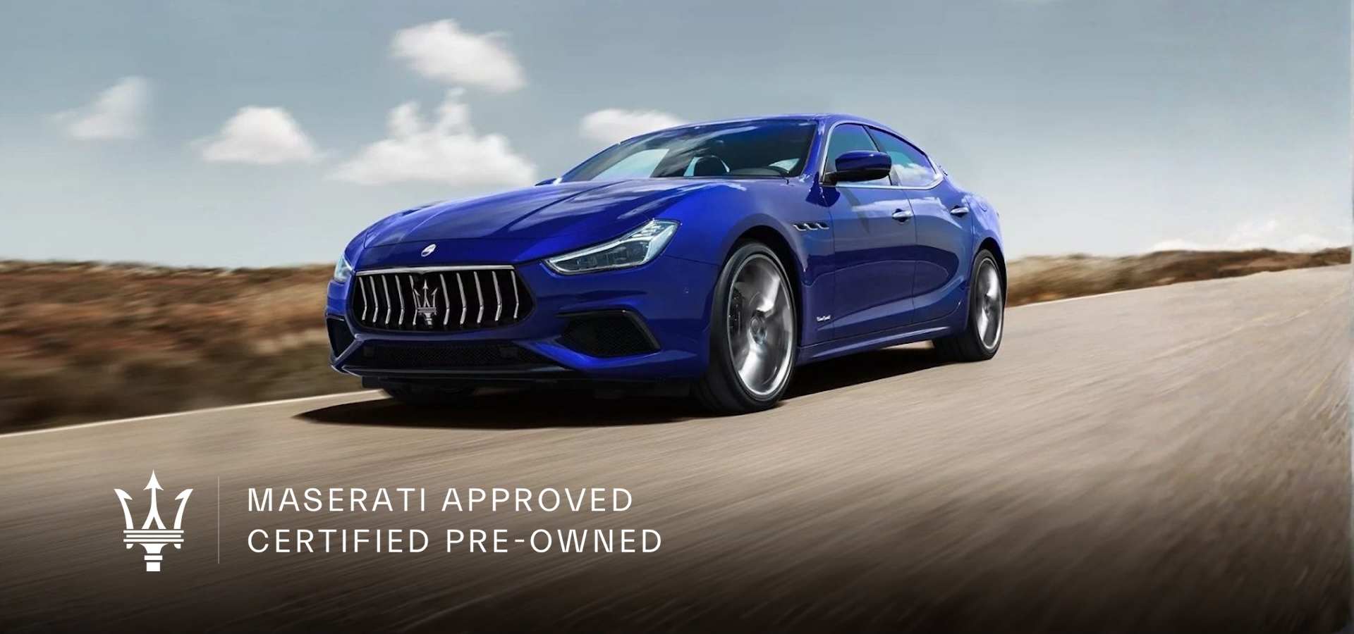 A blue Maserati Ghibli sedan drives along an open desert road under a partly cloudy sky.