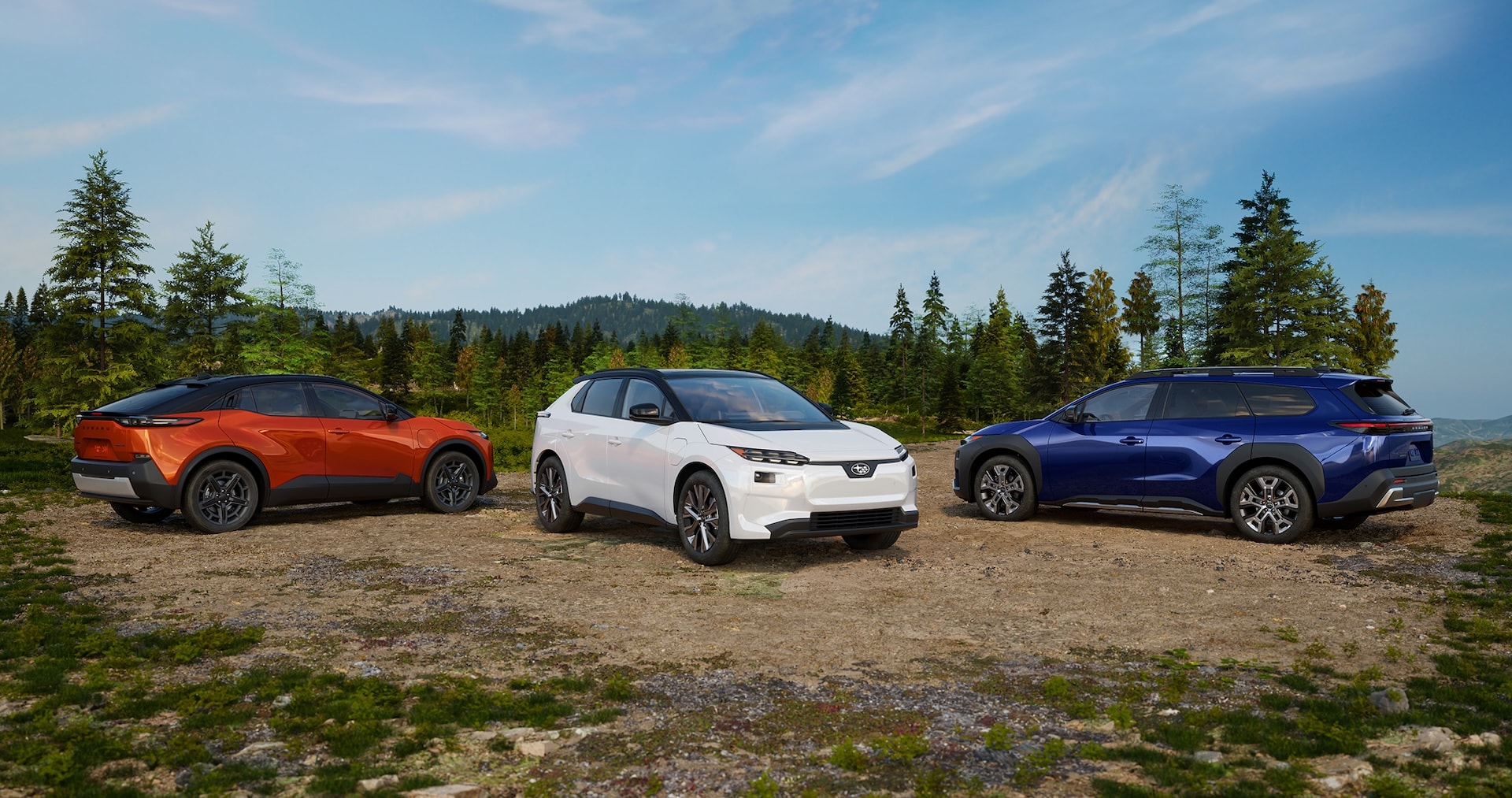 Three Subaru SUVs, one orange, one white, and one blue, are parked on a gravel clearing surrounded by evergreen trees. The vehicles are positioned side by side against a backdrop of rolling forested hills beneath a bright blue sky with wispy clouds. The foreground features rocky, grassy terrain, emphasizing an outdoor and scenic setting.