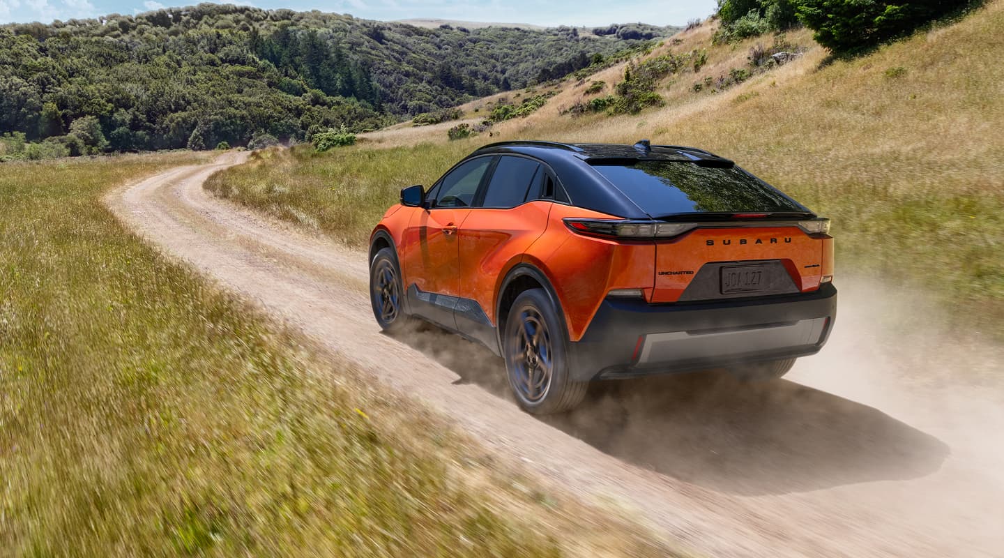 A static image of an orange 2026 Subaru Uncharted driving on a winding dirt road through wide, grassy hills. 