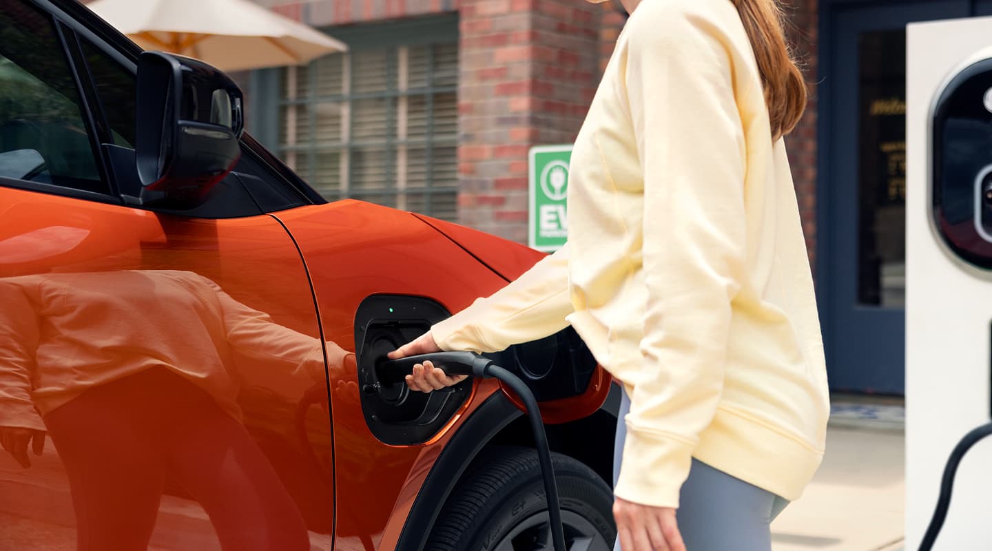 A static close-up image of a person plugging a charging cable into the front driver‑side charging port of an orange Subaru Uncharted. 
