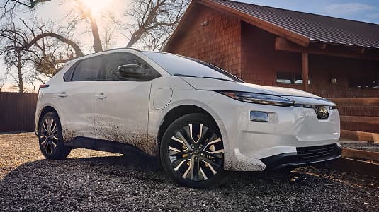 A static image of a white 2026 Subaru Solterra parked on a gravel surface beside a wooden building. 