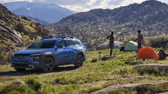 A static image of a blue 2026 Subaru Trailseeker parked on a grassy clearing in a rocky mountain landscape. 