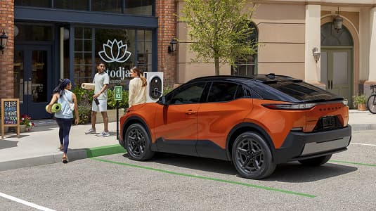 A static image of an orange 2026 Subaru Uncharted parked at an EV charging station along a city street. 