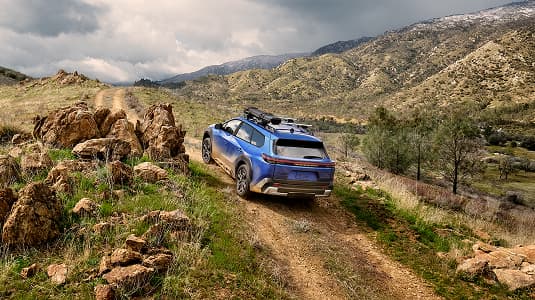 A static image of a blue 2026 Subaru Trailseeker driving on a narrow dirt trail that winds through rocky, grassy hills. 