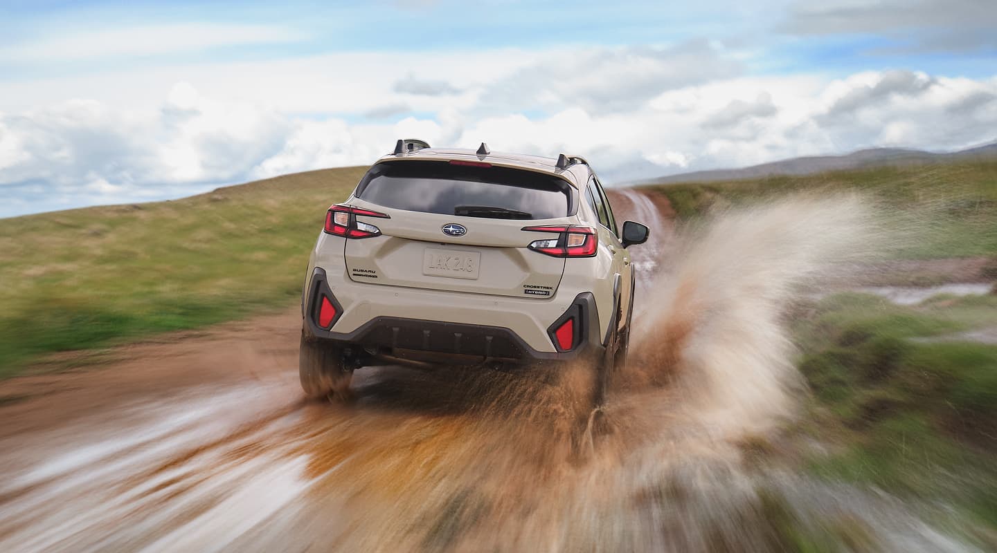 A Subaru Crosstrek Hybrid driving at speed down a muddy trail, kicking up water and mud as it moves through an open landscape under a partly cloudy sky.