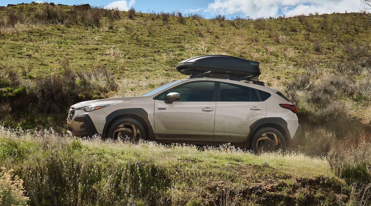 A beige Subaru Crosstrek Hybrid drives off‑road through a grassy, uneven hillside with a rooftop cargo box attached under a bright, partly cloudy sky.