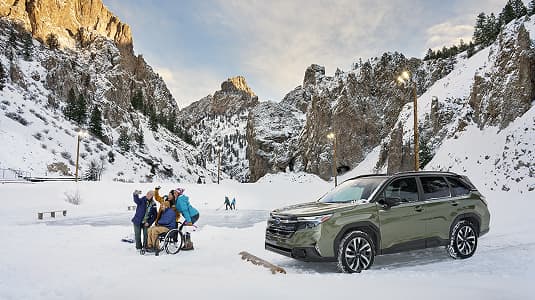 A Subaru Forester Hybrid parked on a snowy mountain road near a group of people gathered around a wheelchair user, with snow-covered cliffs and pine trees in the background under a partly cloudy sky.