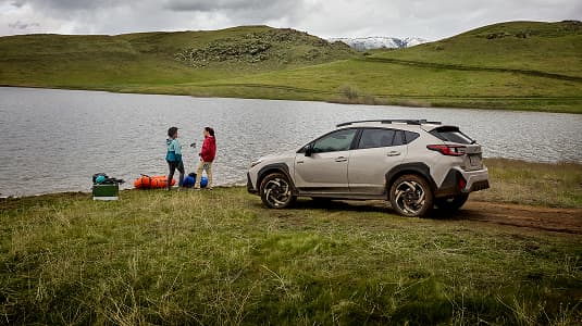 A Subaru Crosstrek Hybrid parked beside a lakeshore on a grassy field, with two people standing near camping gear by the water under a cloudy sky and rolling green hills.