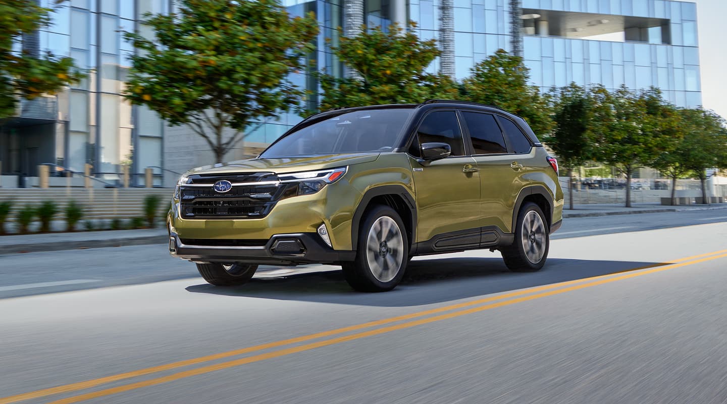 A gold Subaru Forester Hybrid drives down a city street lined with trees and modern glass buildings on a bright day.