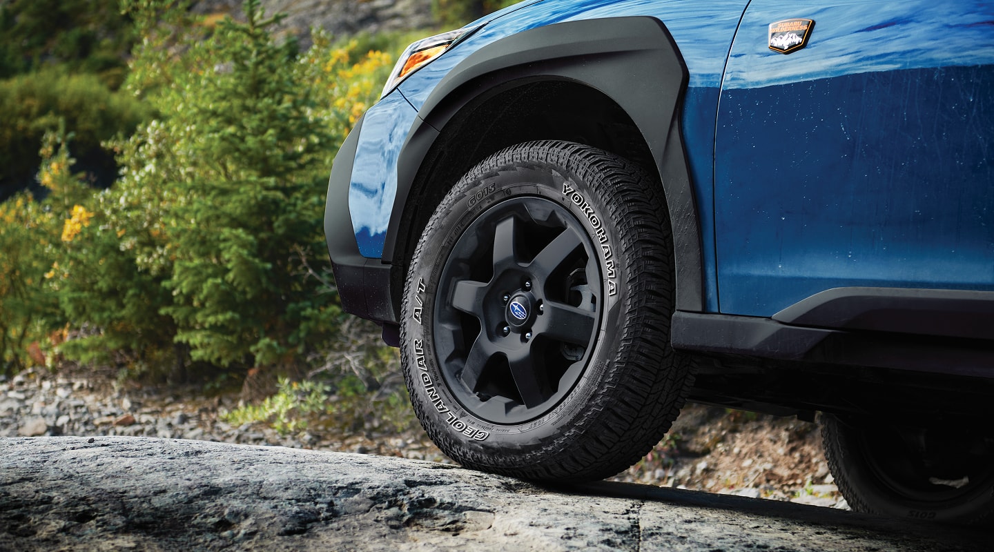 A close-up static image of the front wheel and lower side panel of a blue Subaru Wilderness model parked on uneven rocky terrain. 