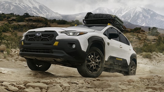 A static image of a 2026 Crosstrek Wilderness driving over a rocky dirt trail in a rugged, mountainous landscape under dark, stormy clouds. 