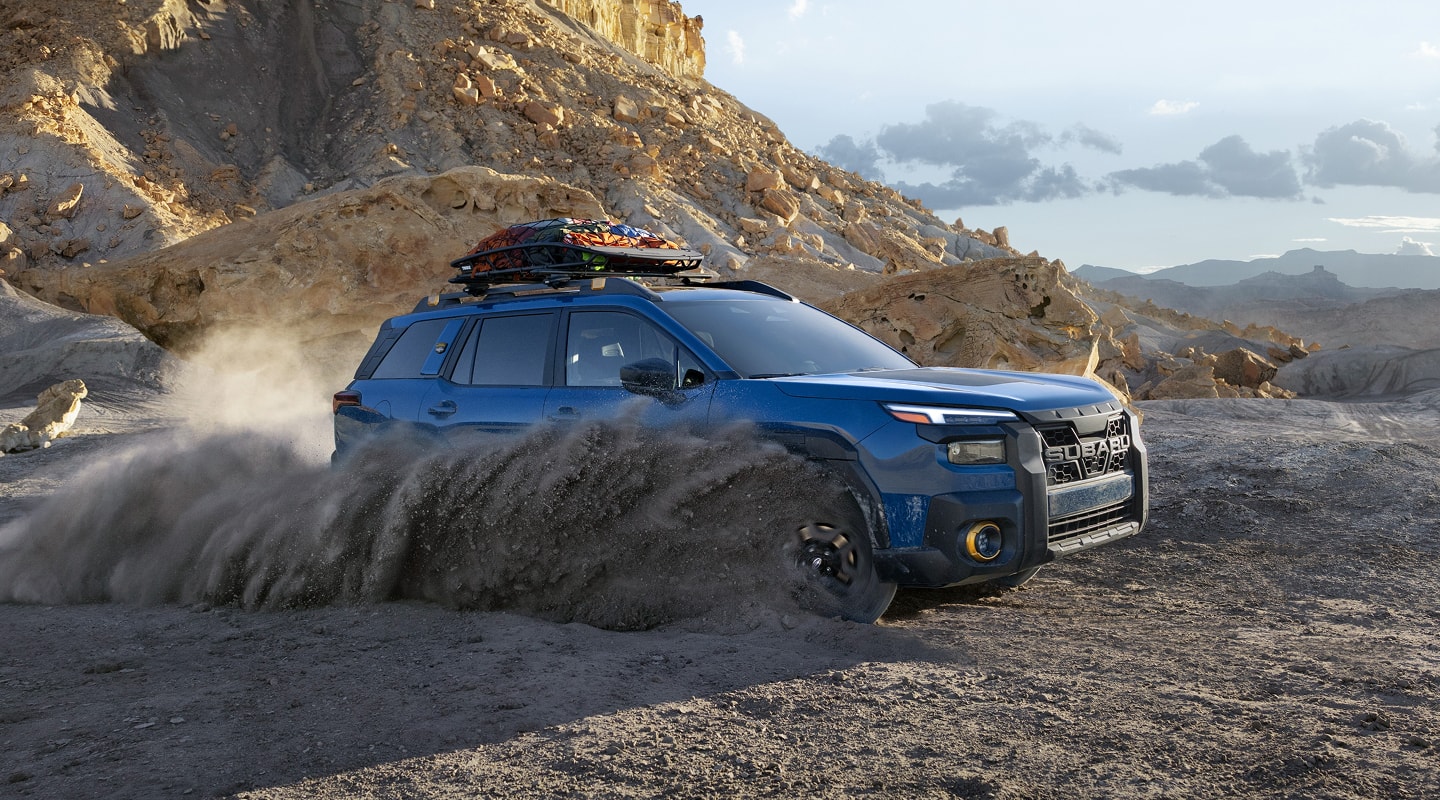 A static image of a blue 2026 Subaru Outback Wilderness driving through loose dirt in a rocky desert landscape. 