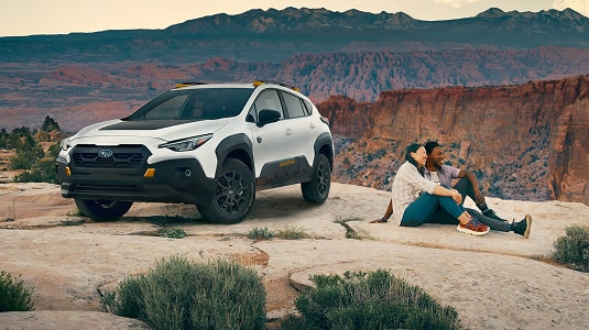 A static image of a white 2026 Subaru Wilderness model parked on a flat rock surface overlooking a wide desert canyon.