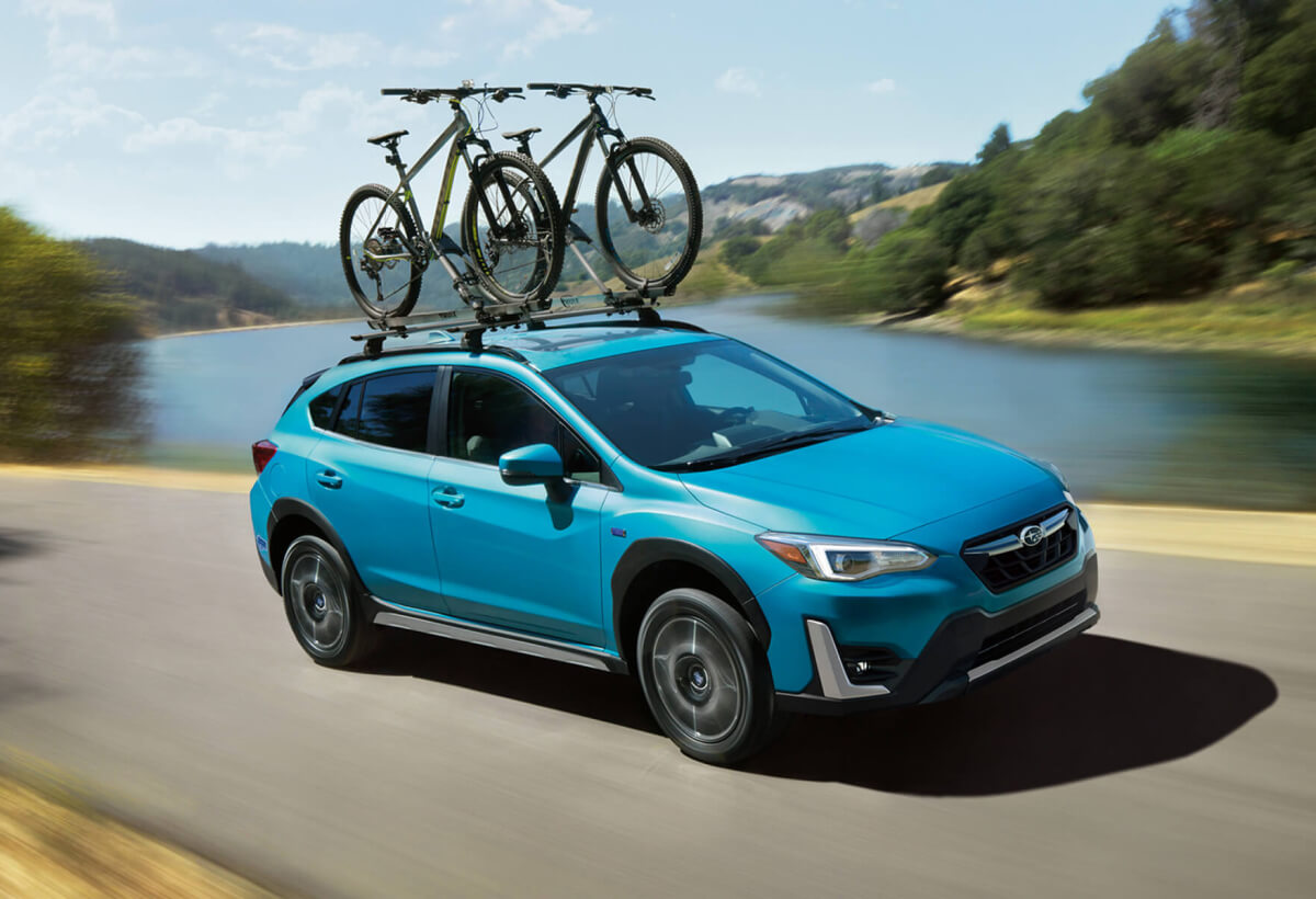 A blue Crosstrek Hybrid with two bicycles on its roof rack driving beside a river