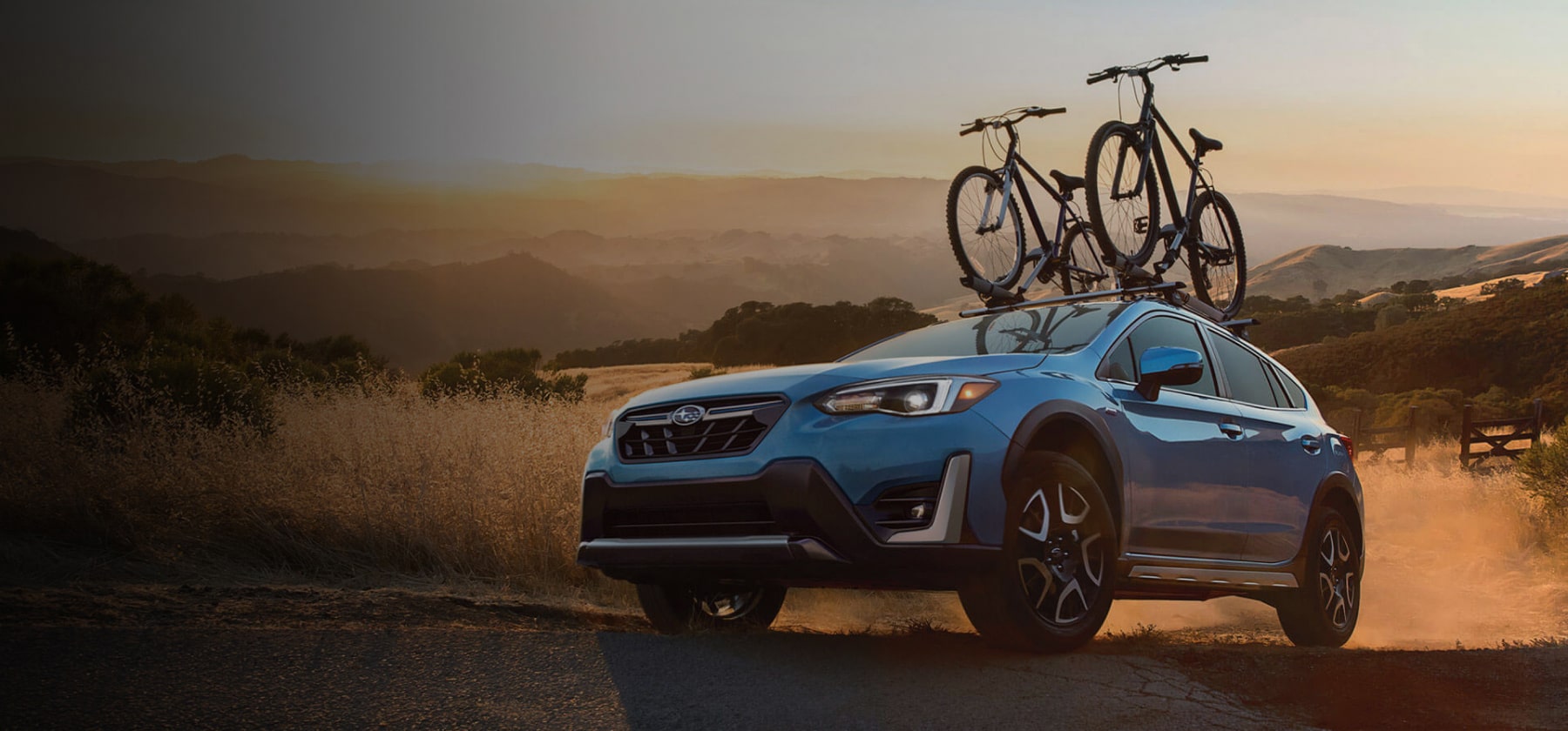 A blue Crosstrek Hybrid with two bicycles on its roof rack parked in front of low mountains