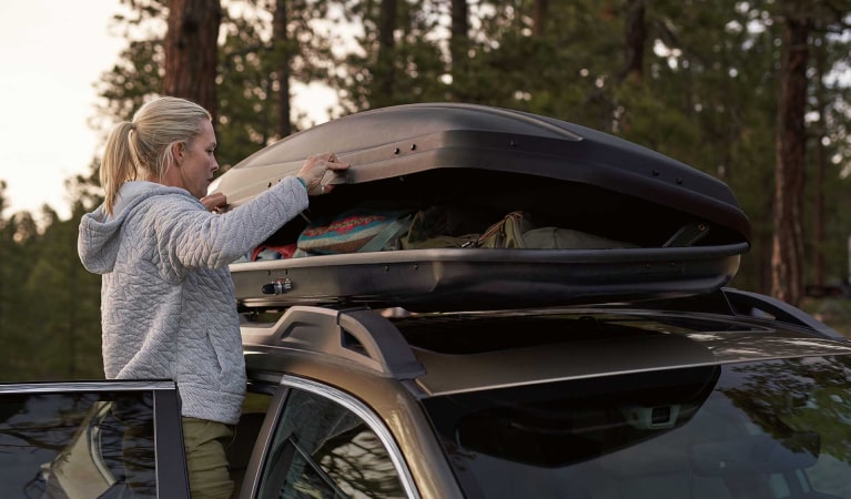 A woman opening a cargo container accessory mounted to the standard raised roof rails on a 2022 Subaru Outback.