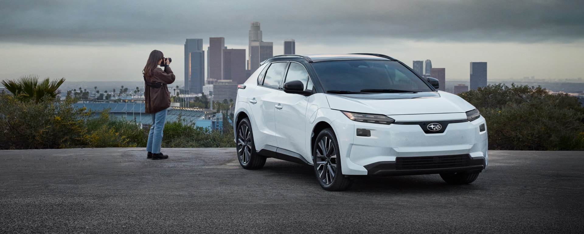 A person takes a photo next to a white Solterra EV parked in front of a city skyline.
