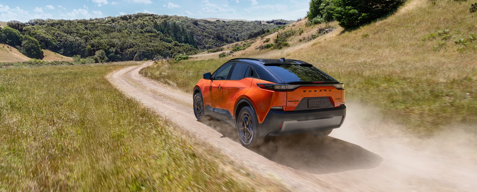 An orange Subaru Uncharted electric SUV drives on a dirt road through some rolling hills.