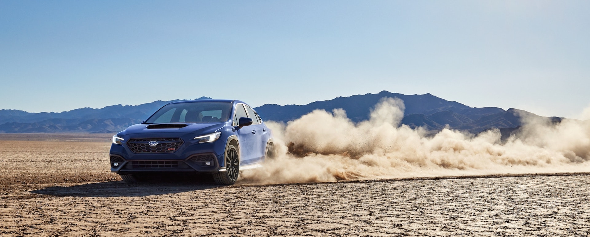 A WRX high-performance sports sedan drifts a turn on a desert bed, kicking up a cloud of dust.