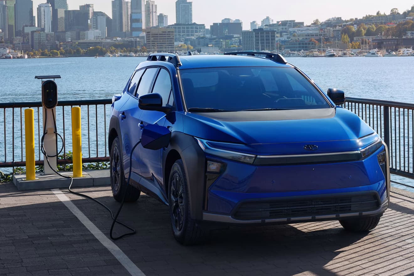 Blue 2027 Toyota bZ Woodland parked beside a waterfront with a city skyline behind it