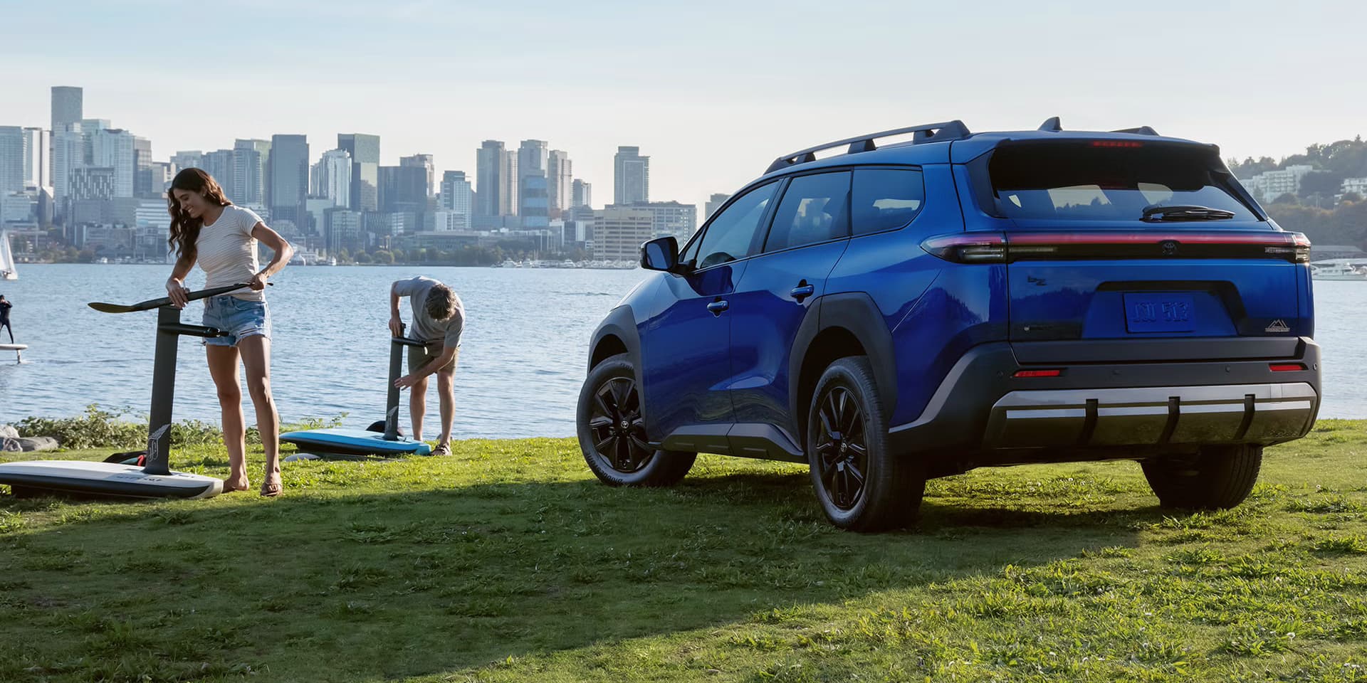 Blue 2027 Toyota bZ Woodland parked on a grassy waterfront with a city skyline and people paddleboarding