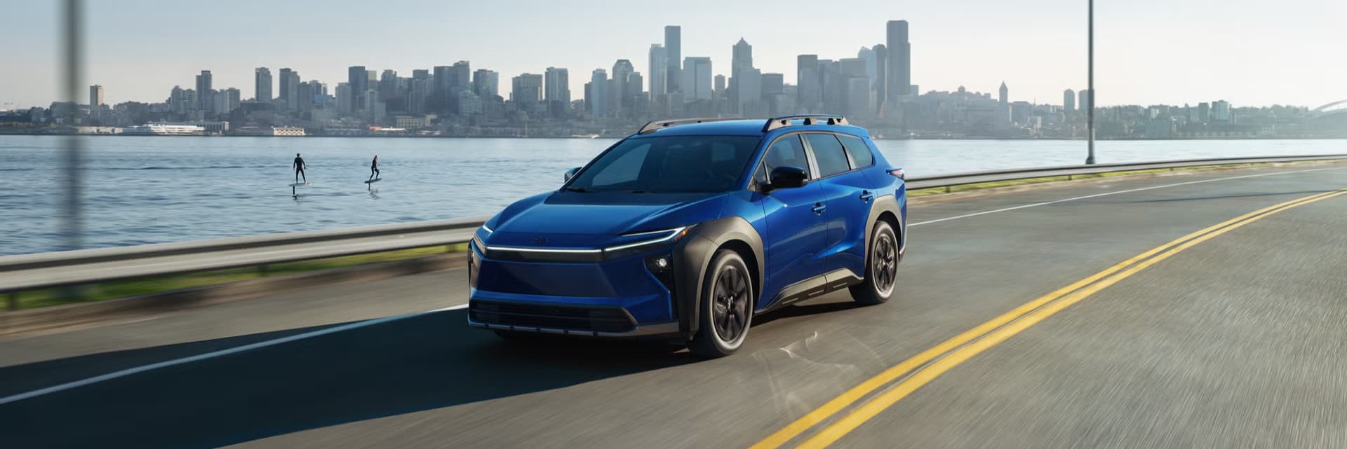 Blue 2027 Toyota bZ Woodland driving along a waterfront road with a city skyline behind it